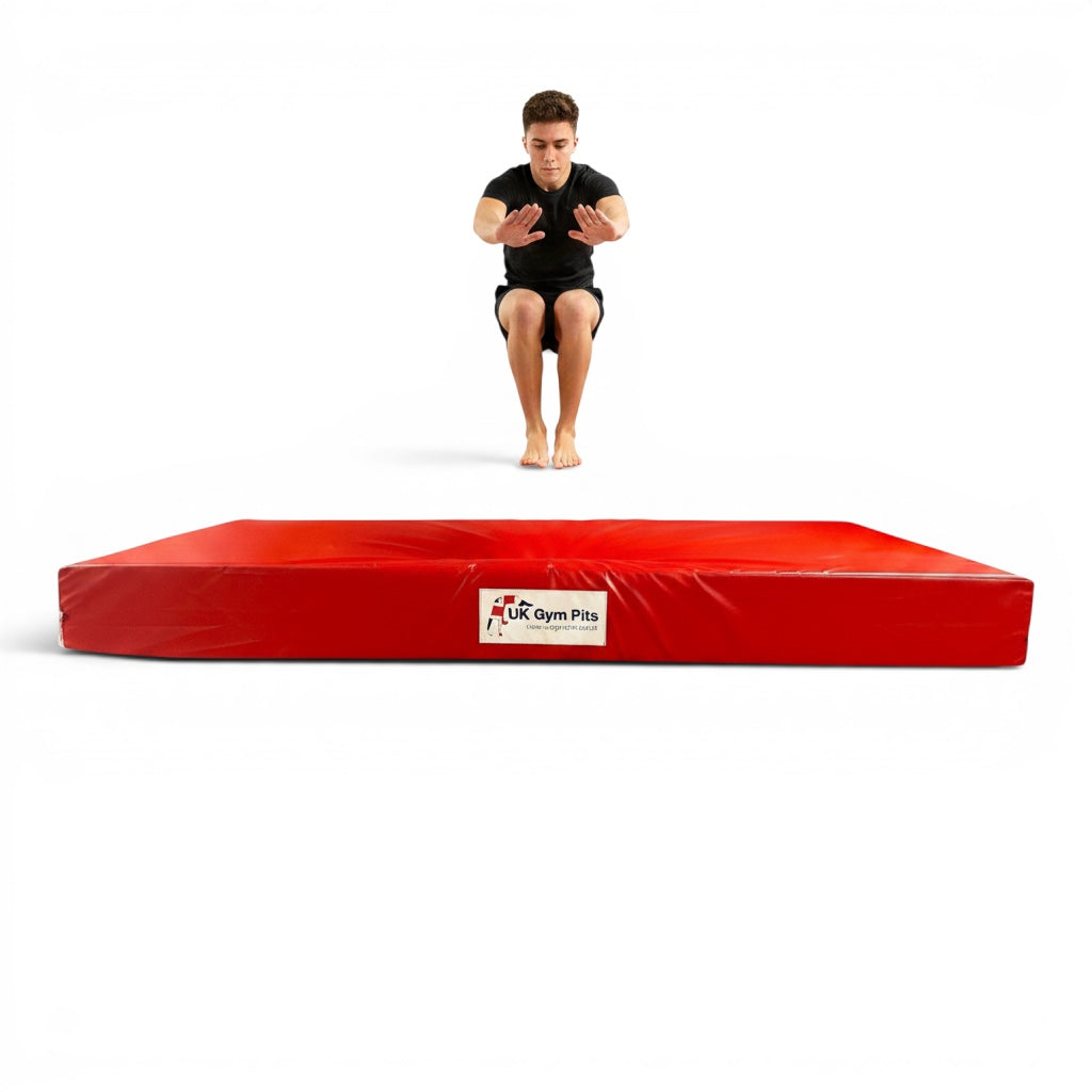 UK Gym Pits red crash mat with a gymnast jumping above it, featuring shock-absorbing foam and heavy-duty PVC cover.